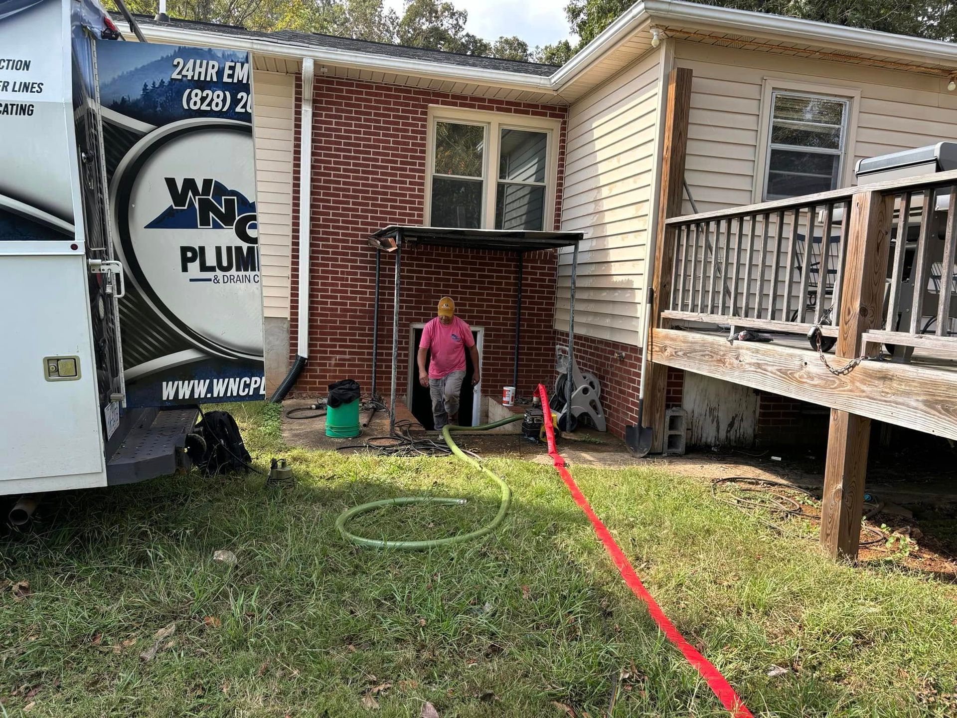 Person stands by a brick house with a garden hose and a red extension cord near a raised wooden deck.