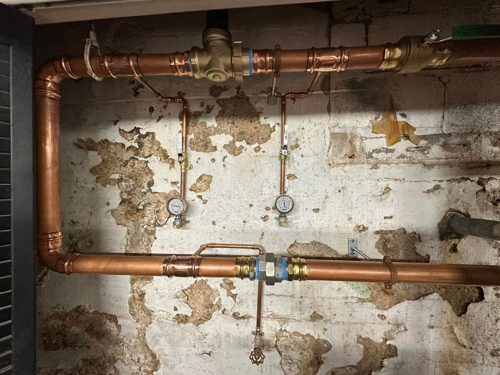 Old basement wall with exposed copper pipes and peeling paint above a rough concrete floor