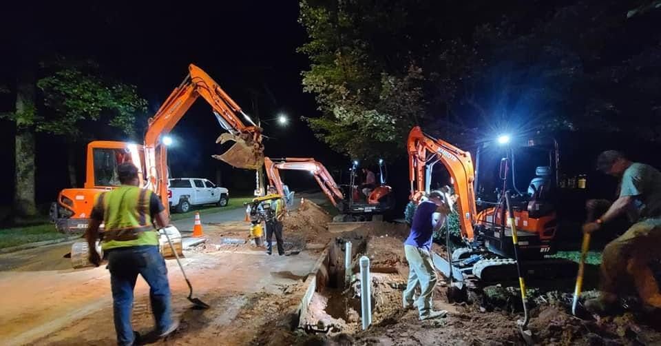 Nighttime roadwork with excavators and workers digging under bright lights along a street.