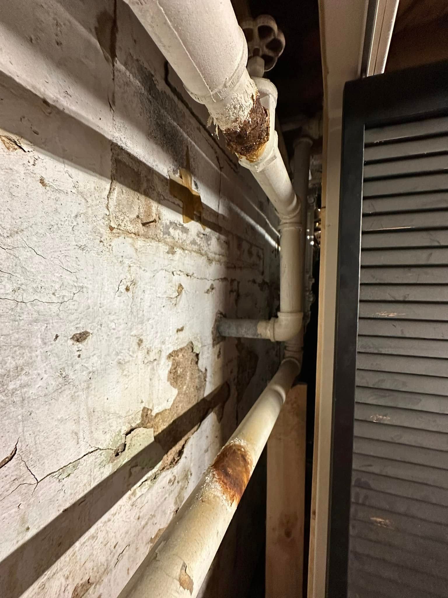 Rusty white pipes along a rough wall beside a dark louvered panel.