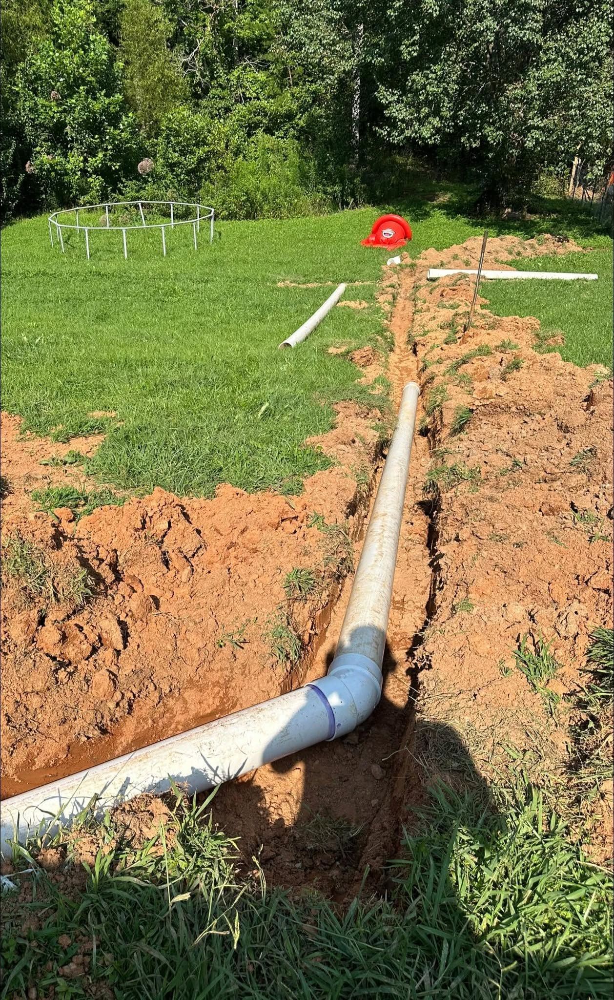 White irrigation pipe running through a trench in a grassy field near trees, with a red marker in the background