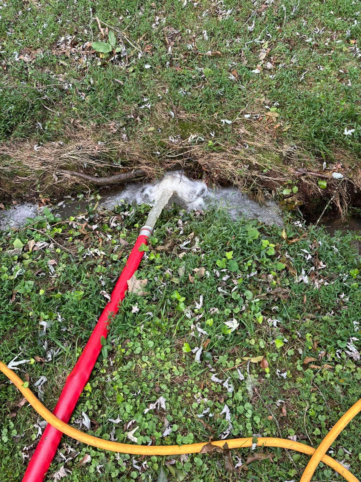 Red hose spraying water across a grassy yard with a small ditch or stream.