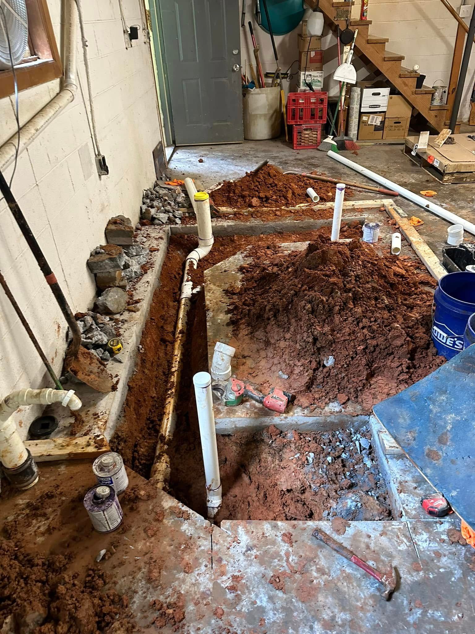Indoor plumbing excavation with exposed pipes, dirt pile, and tools in a narrow work area