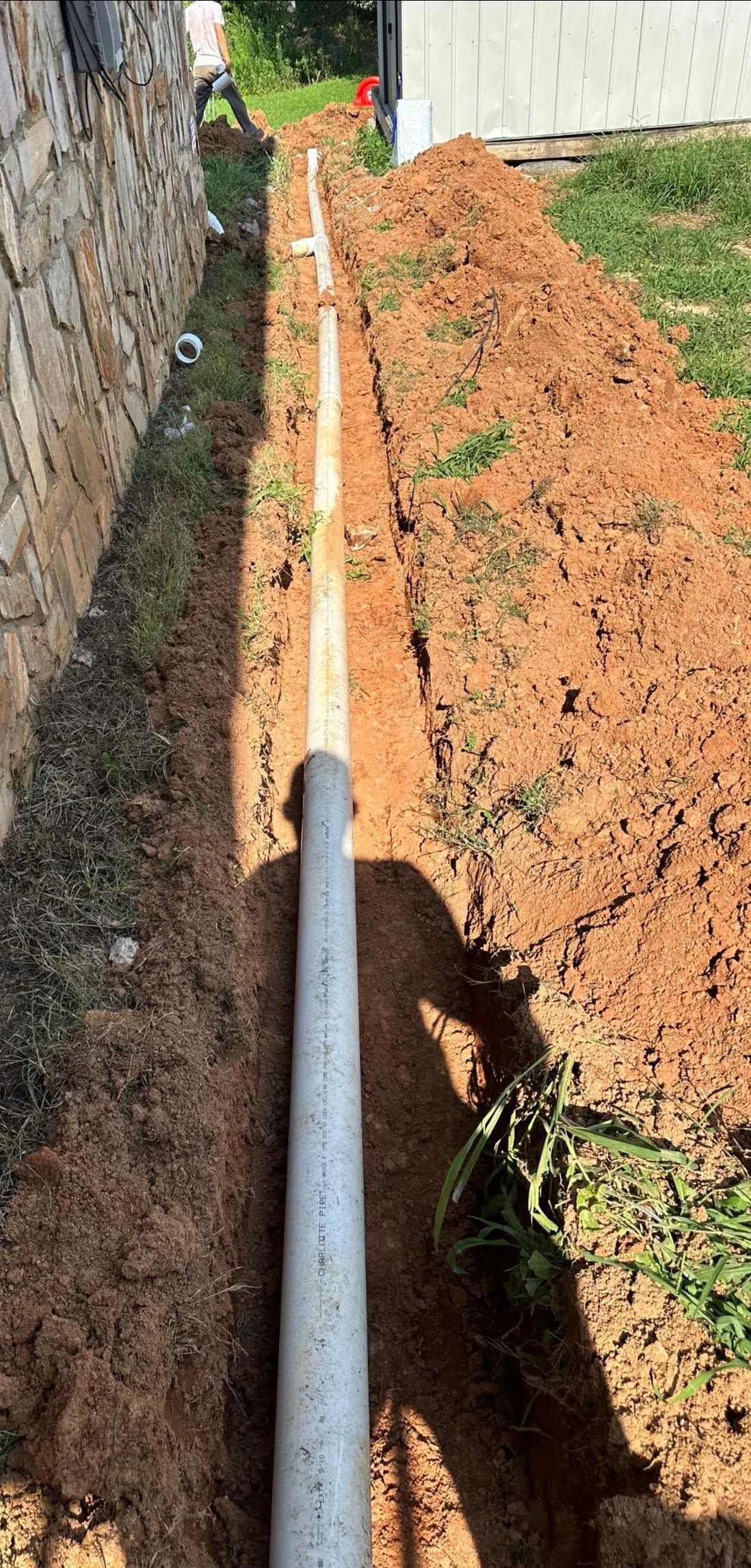 White pipe laid in a narrow trench beside a stone wall, with orange dirt and grass along the edge.