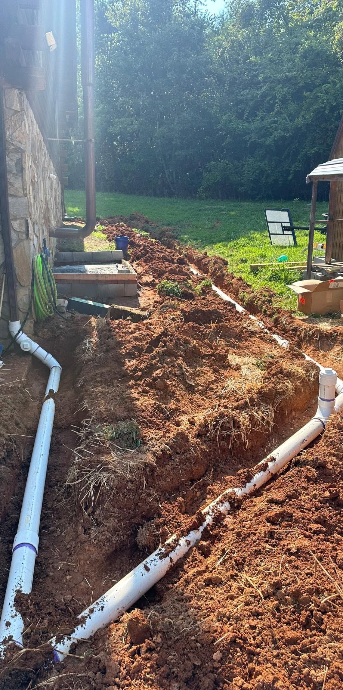 Sunlit garden path with white pipes, mulch, and raised beds beside a stone wall.