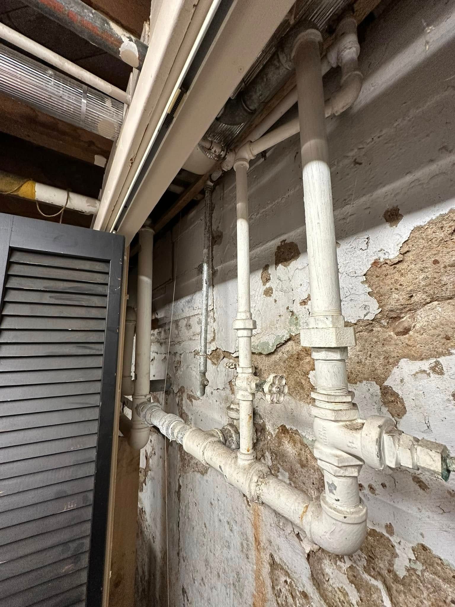 Old, rusted plumbing pipes along a peeling wall in a cramped utility space
