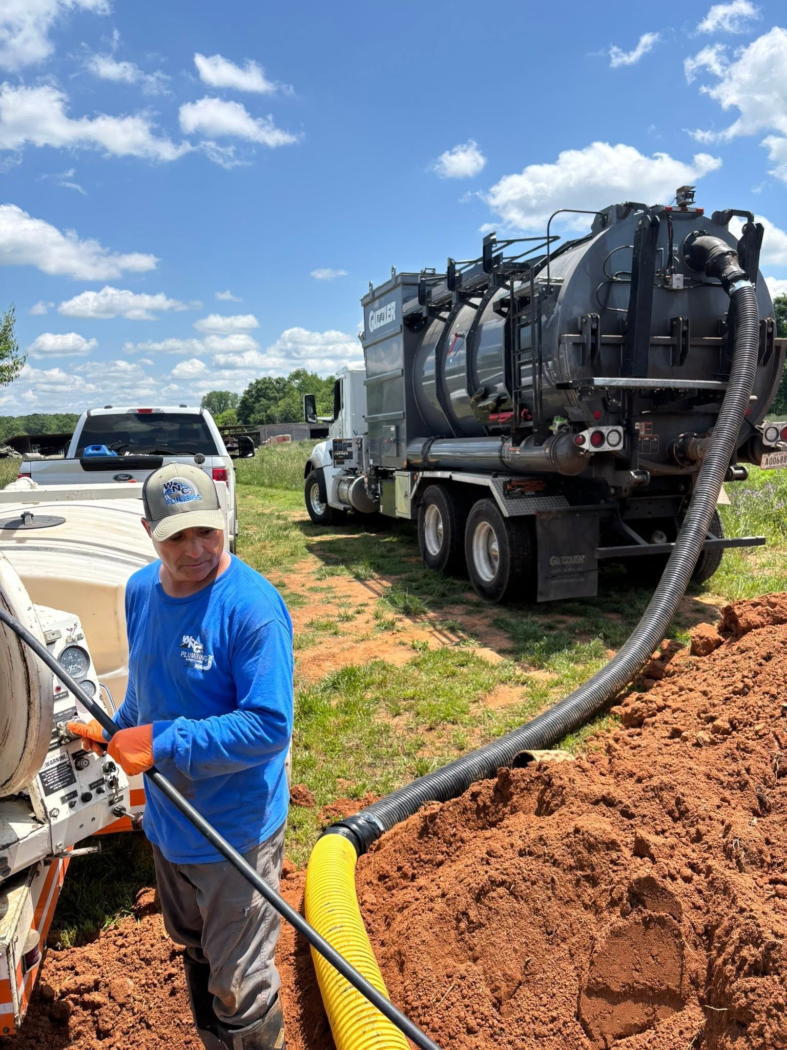 Worker in blue shirt operating hoses beside a vacuum truck in a muddy field under a bright sky.