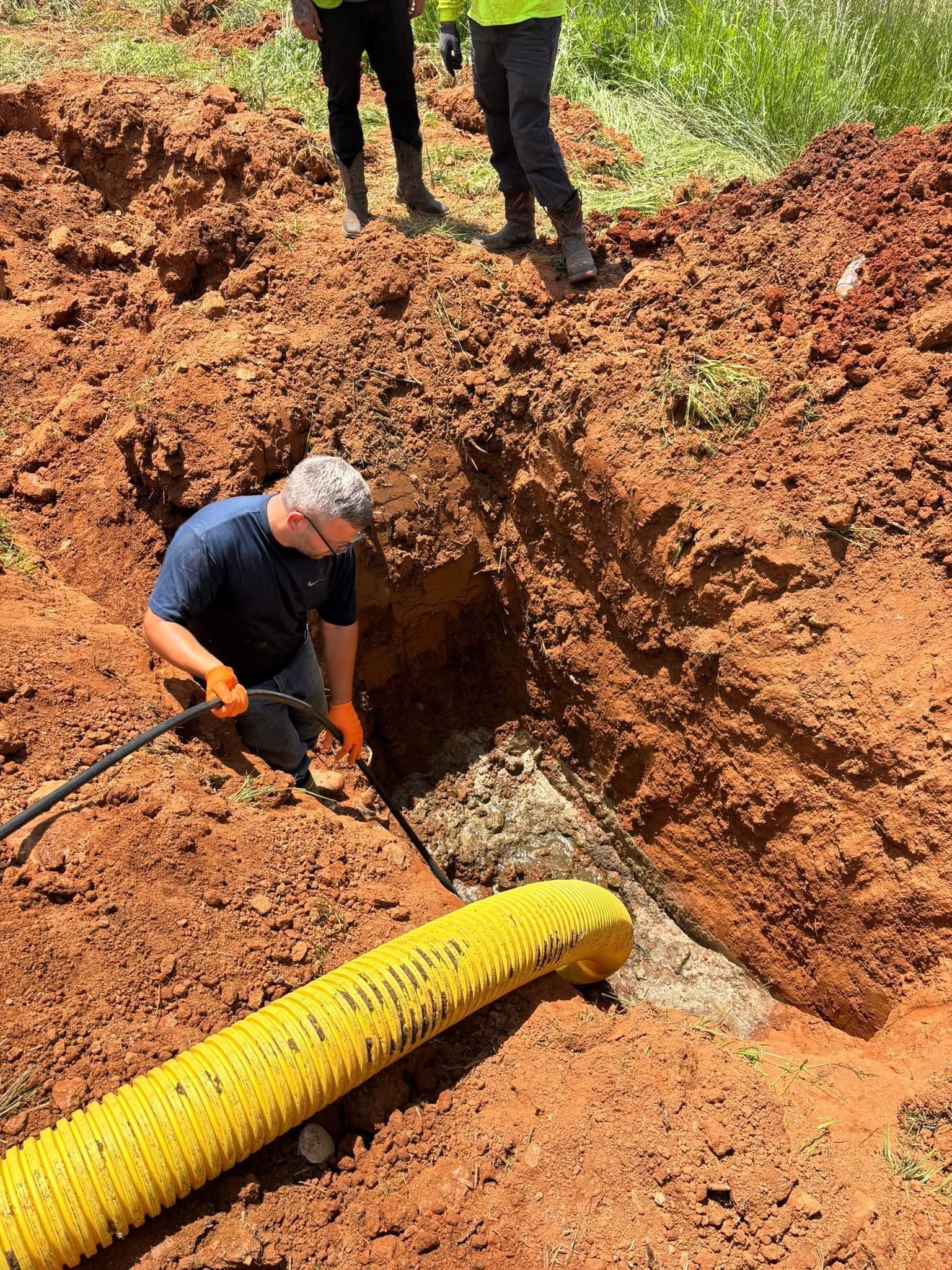 Worker excavating a trench beside a yellow pipe in a red dirt construction site