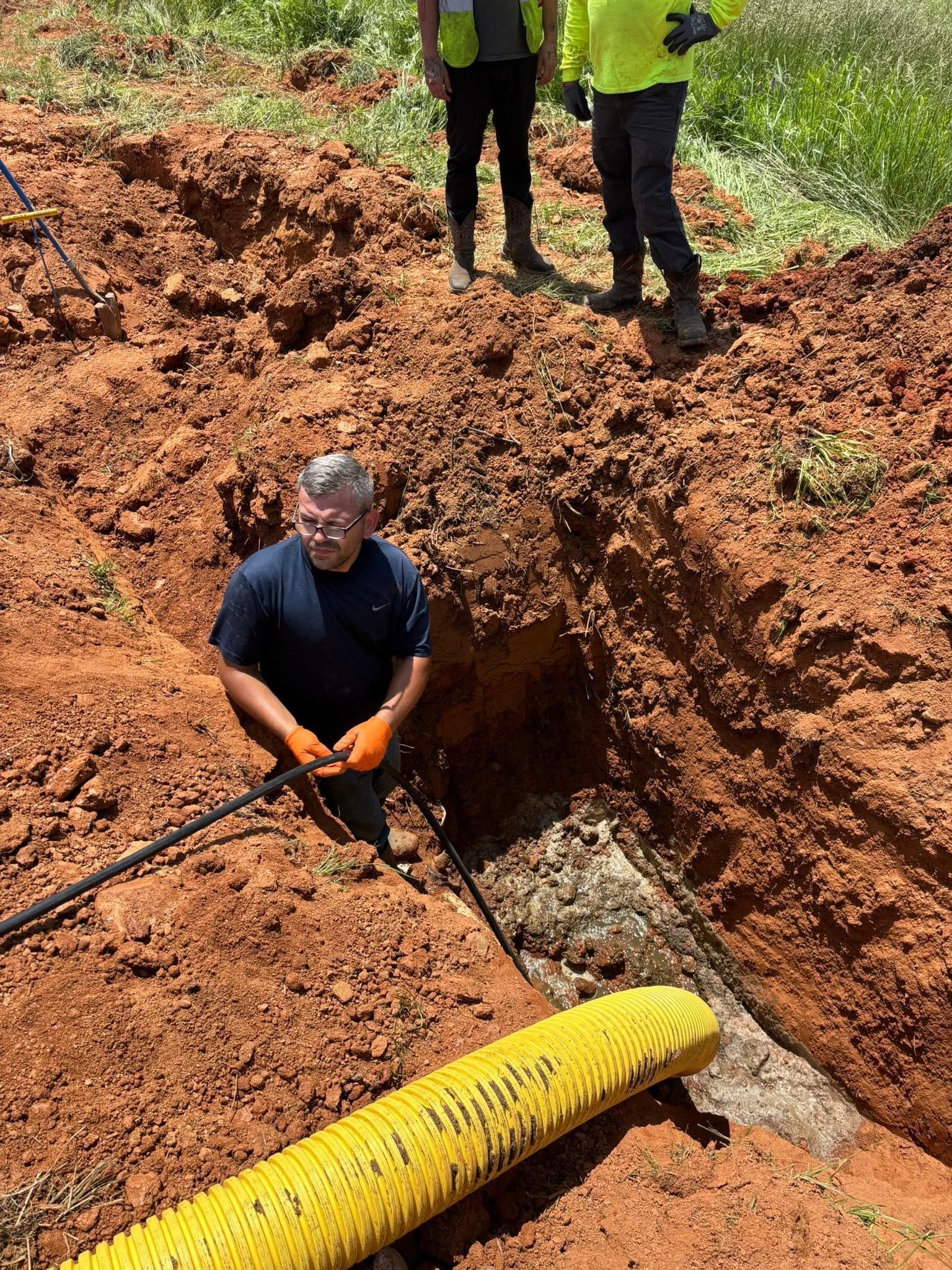 Workers excavating a muddy trench near a yellow drainage pipe on a hillside