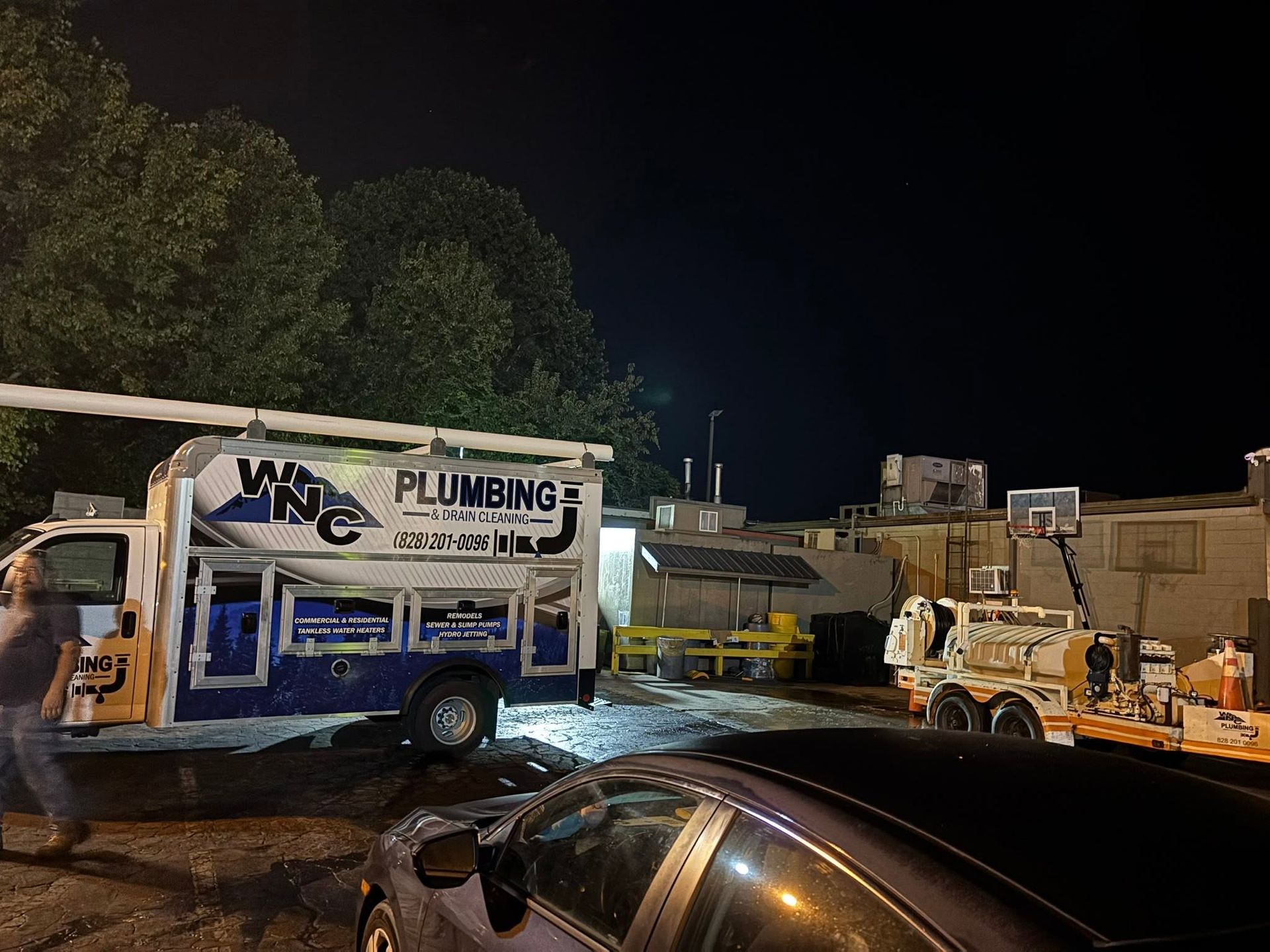 Plumbing service truck and excavator at a nighttime work site with parked car in foreground