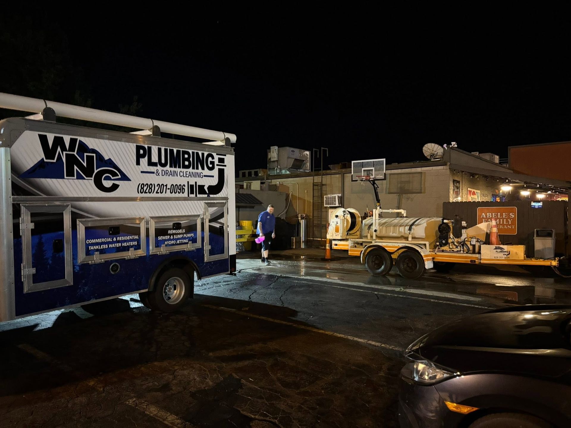Plumbing service truck at night outside a building with a trailer and equipment lit by work lights