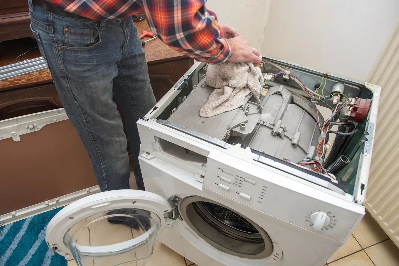 A person in a plaid shirt repairs a washing machine with an exposed top, using a cloth to wipe down the drum.