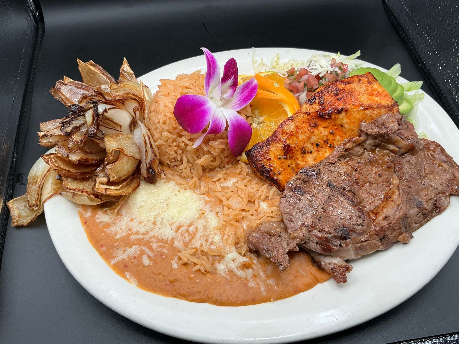 Plate of food: steak, salmon, rice, beans, onion rings, orange pepper, garnished with a flower.