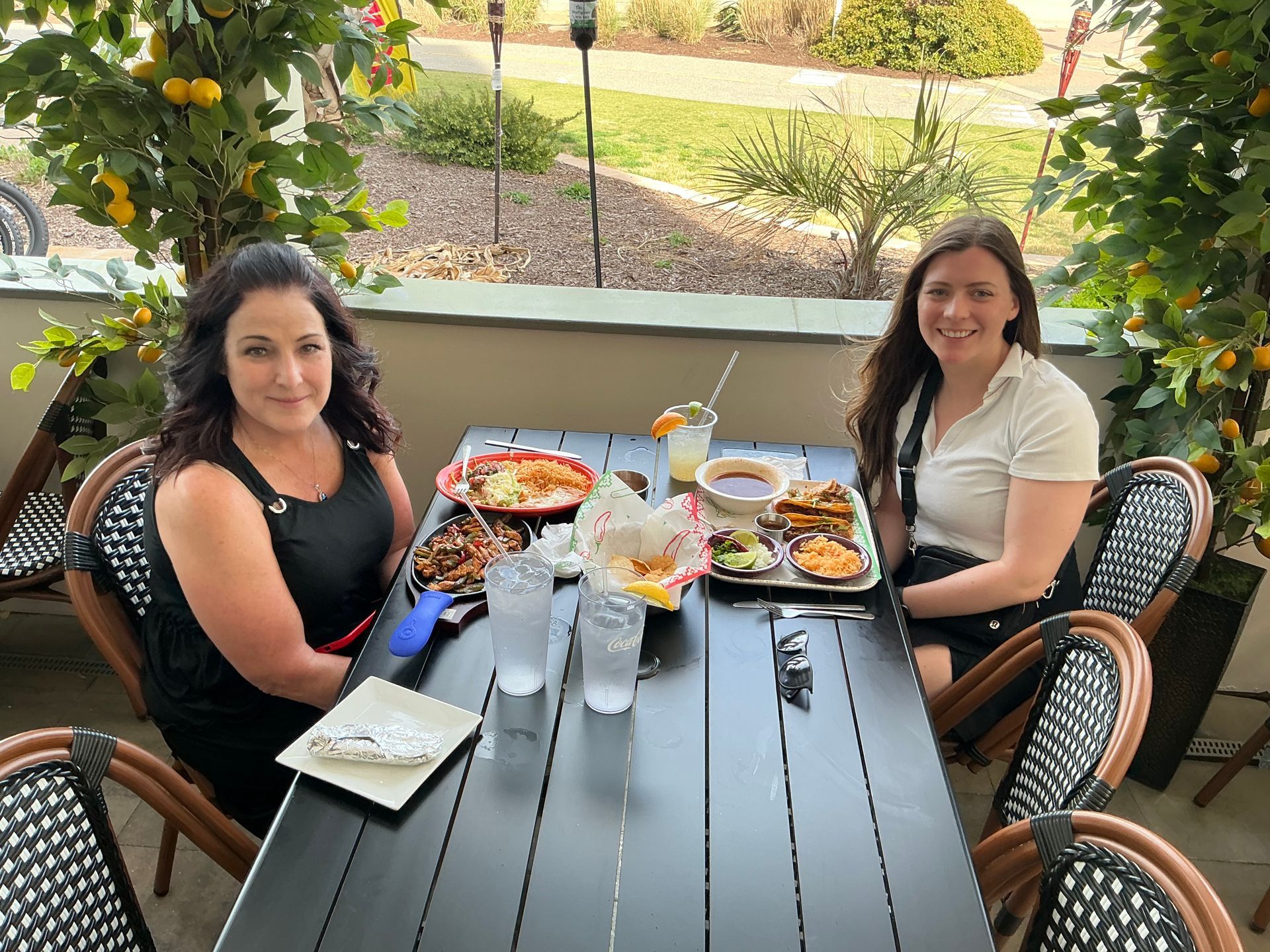 Two people sitting at a table with food and drinks outdoors, smiling.