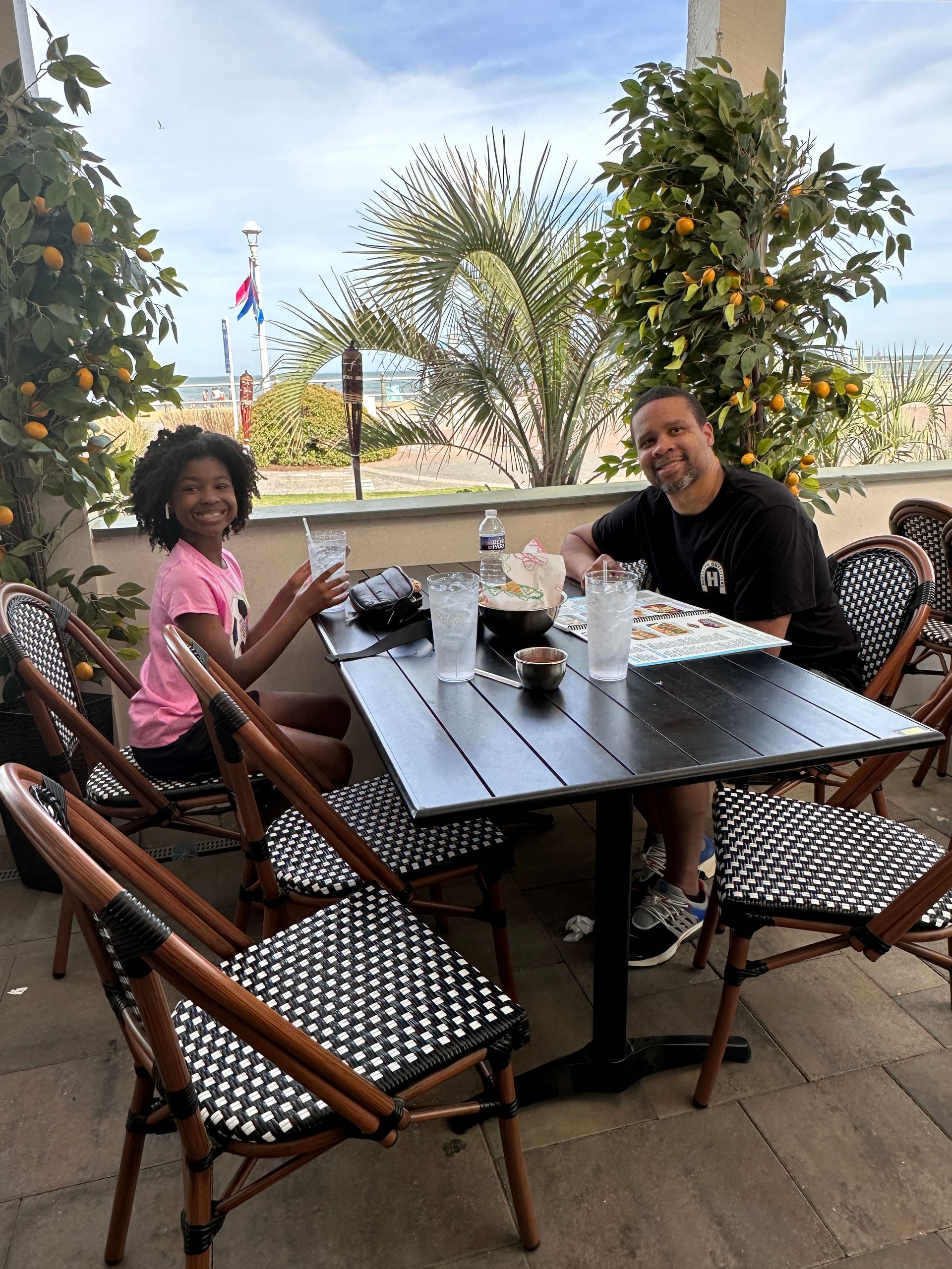 A person and child seated at an outdoor restaurant table. They eat food, looking at camera. Beach in background.
