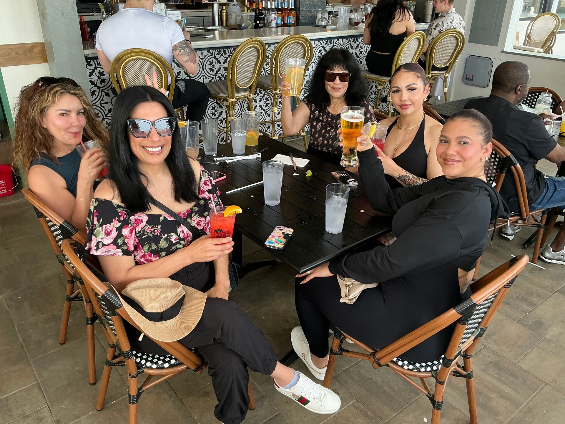 Five people at an outdoor table, toasting drinks. Smiling, relaxed atmosphere. Bar and people in background.