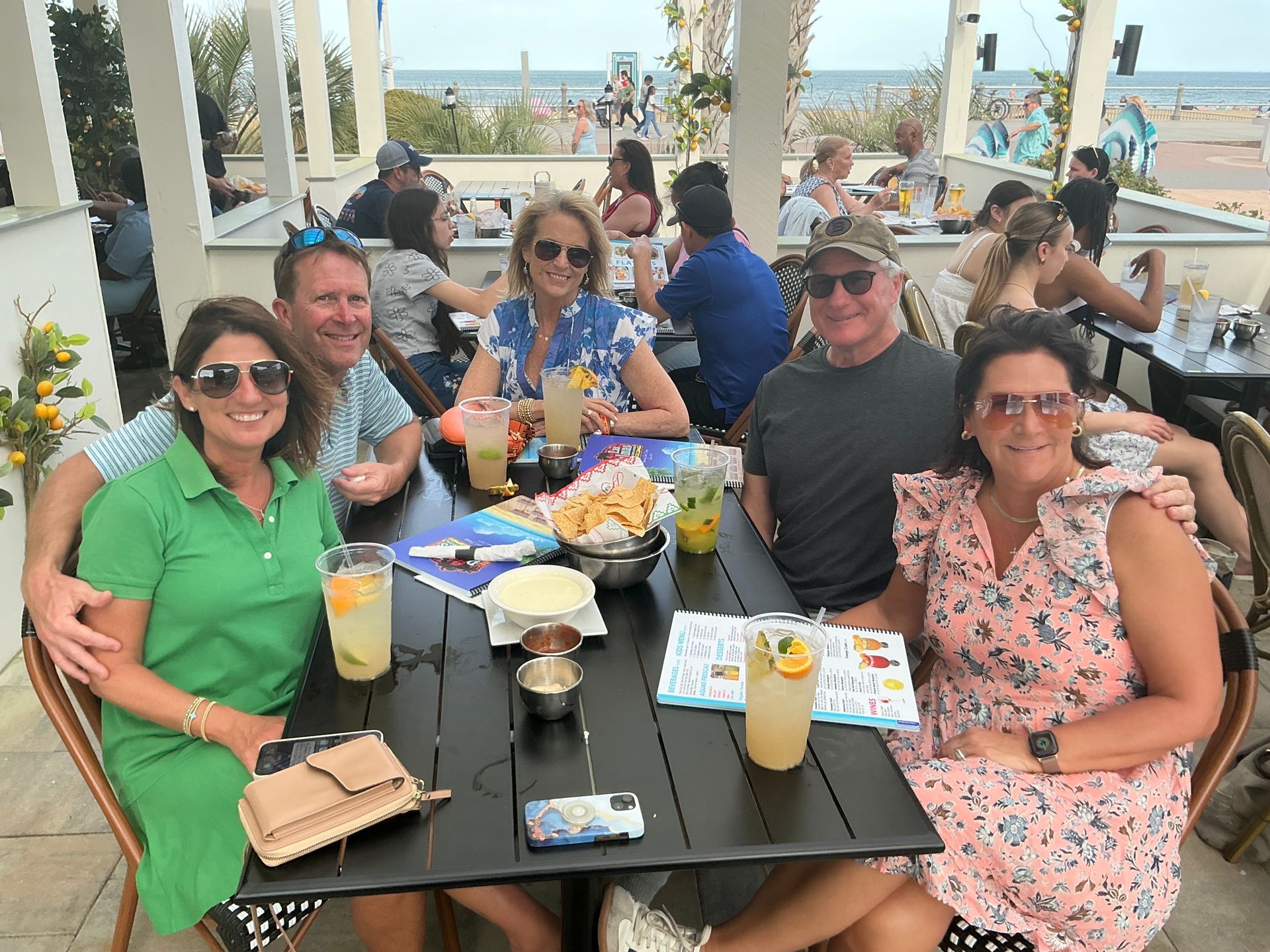 Group of people at an outdoor restaurant, ocean view. They are smiling and eating at a table with food and drinks.