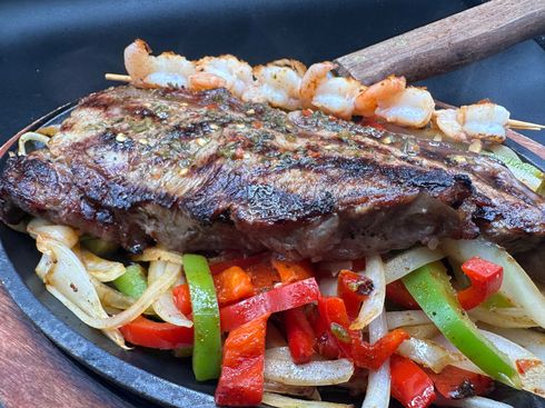 Sizzling steak and shrimp skewers atop sauteed peppers and onions on a dark platter.