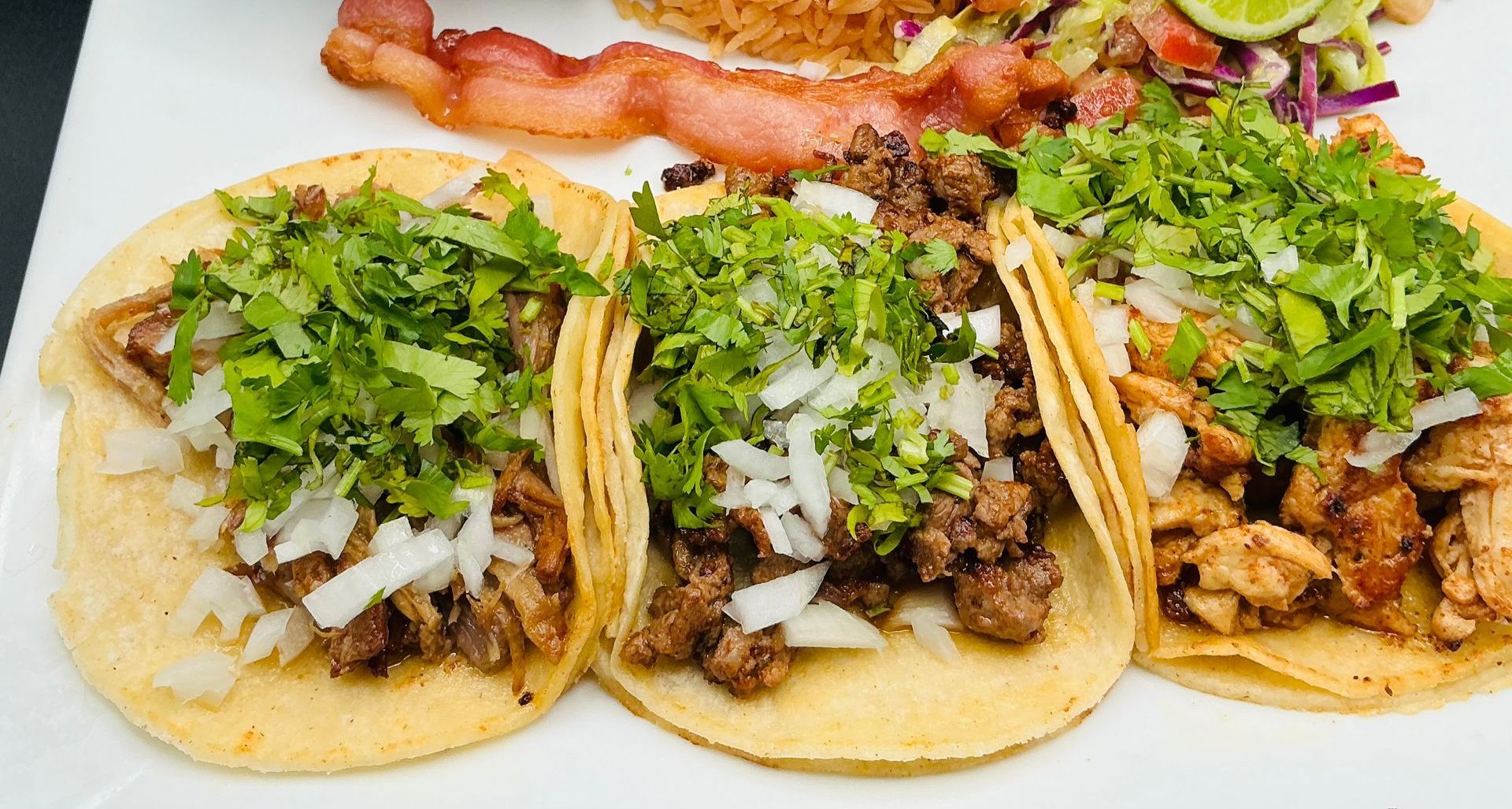 Three tacos topped with cilantro and onions, with bacon and salad in the background.