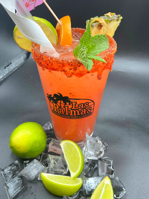 Red cocktail in a cup with lime and pineapple garnish, surrounded by ice and limes.