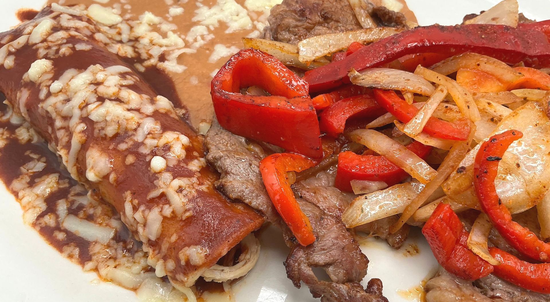 Enchiladas, refried beans, and steak fajitas with red peppers and onions on a plate.