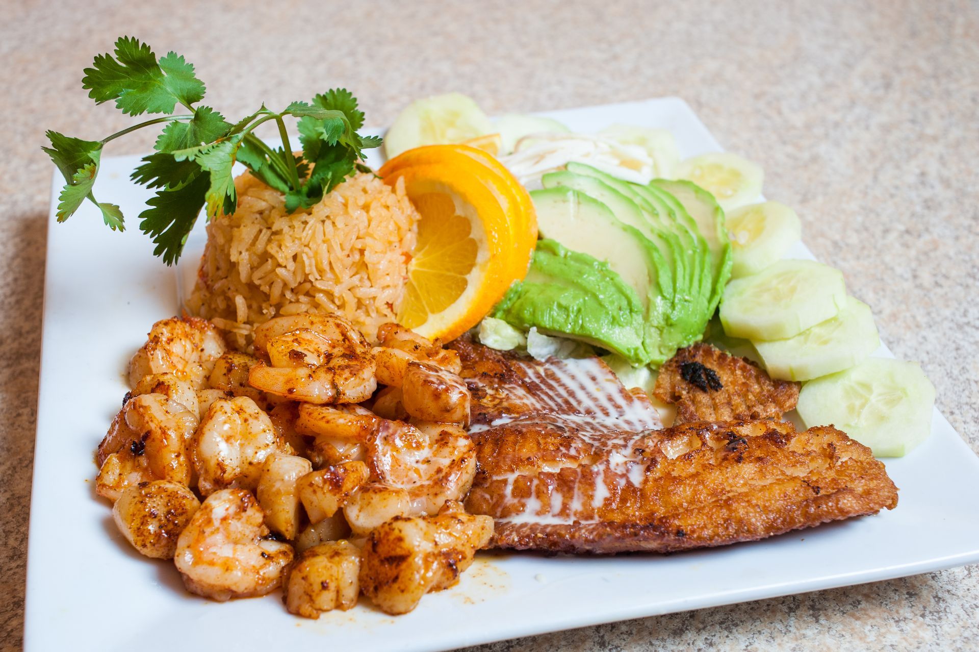 Plate of Mexican-style food with rice, grilled fish, seasoned shrimp, avocado, cucumber, orange slice, and cilantro.