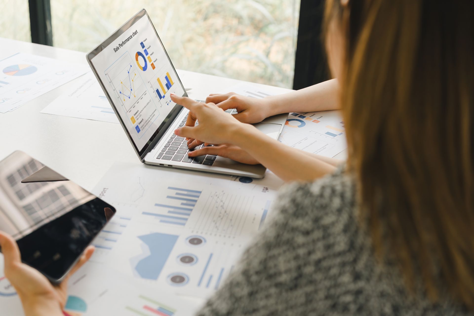 Hands working on a laptop with charts and graphs spread across a desk