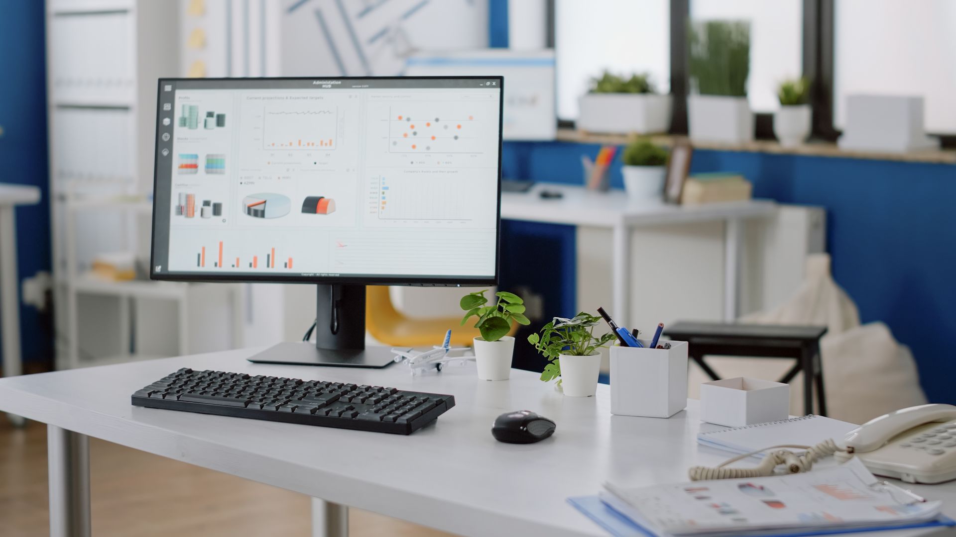 Modern office desk with desktop monitor, keyboard, mouse, notebook, and small plants.
