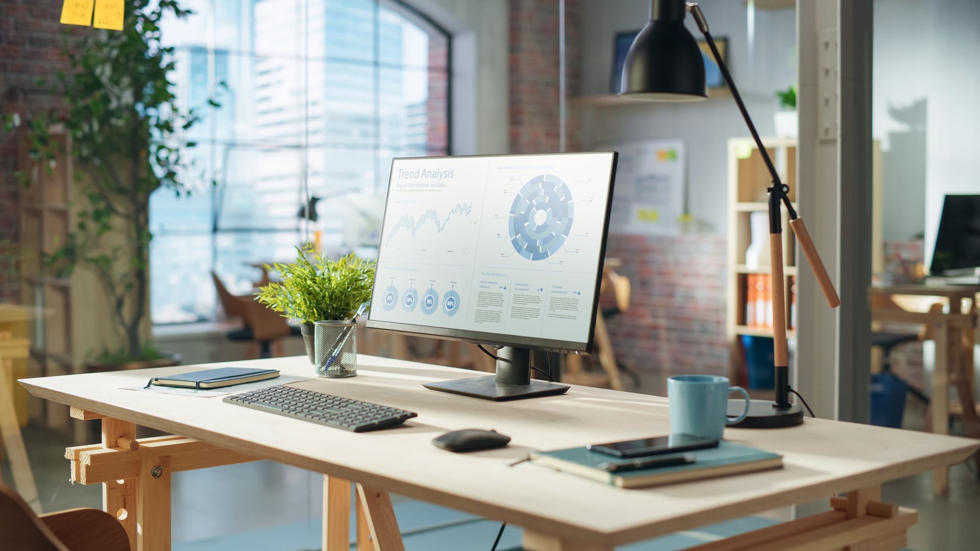 Modern desk workspace with laptop, monitor showing charts, keyboard, plants, and a desk lamp by a window