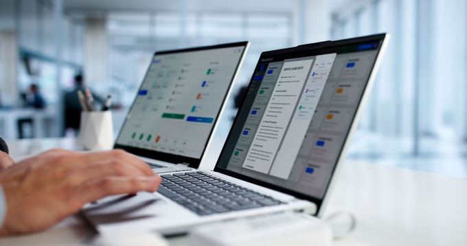 Hands typing on a laptop with two bright screens showing app dashboards in an office setting