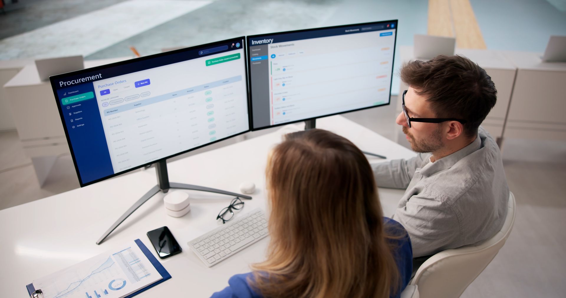 Two coworkers review data on dual monitors at a modern white desk