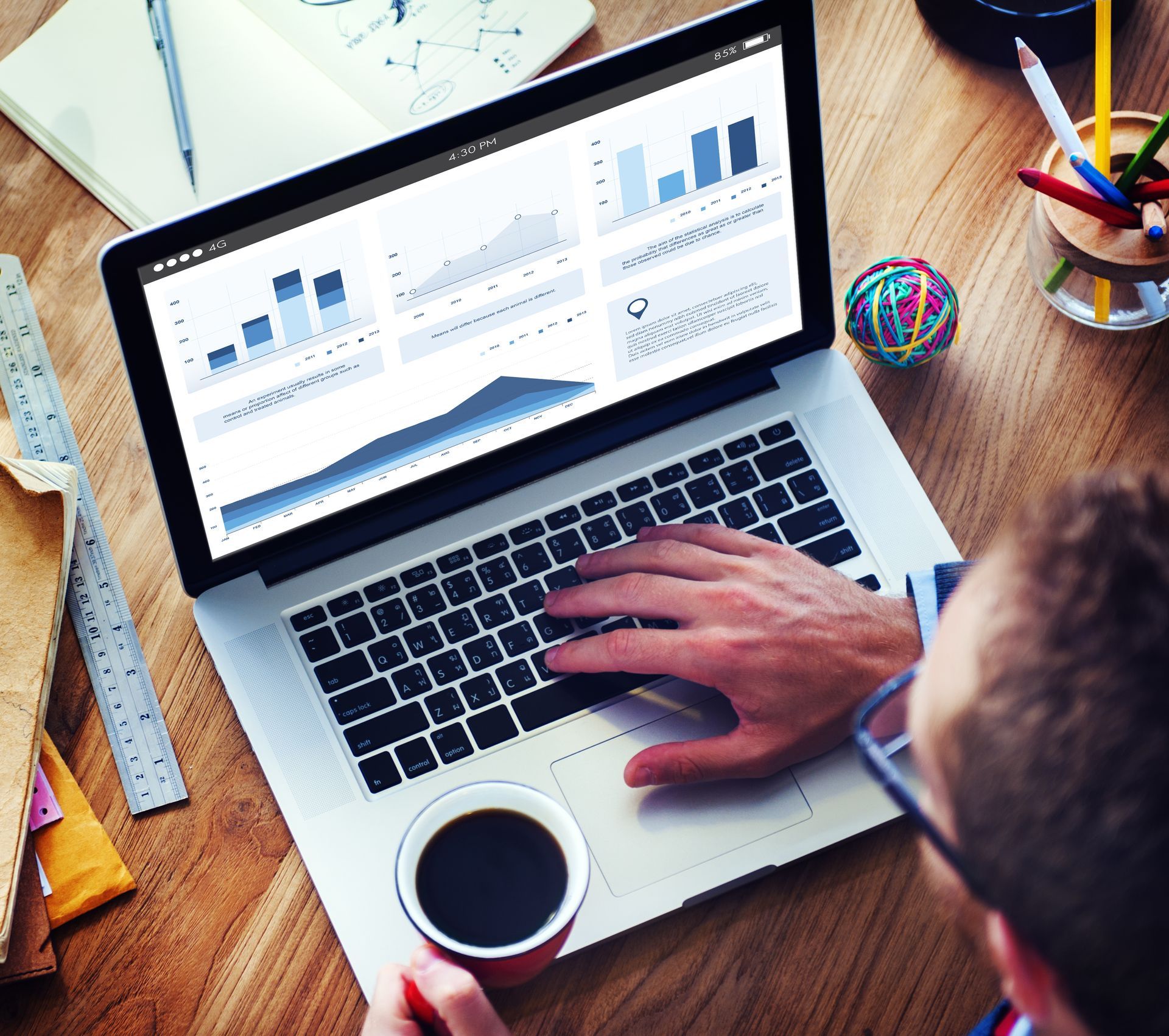 Person working on a laptop with charts and graphs, holding a coffee cup at a wooden desk.