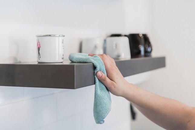 Hand wiping a dark shelf with a blue cloth; mugs are on the shelf.