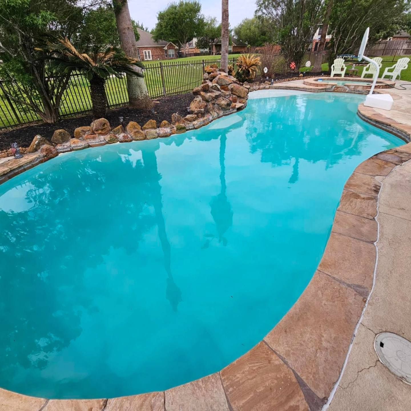 Curved backyard swimming pool with bright blue water, stone deck, and lawn chairs by a fence.