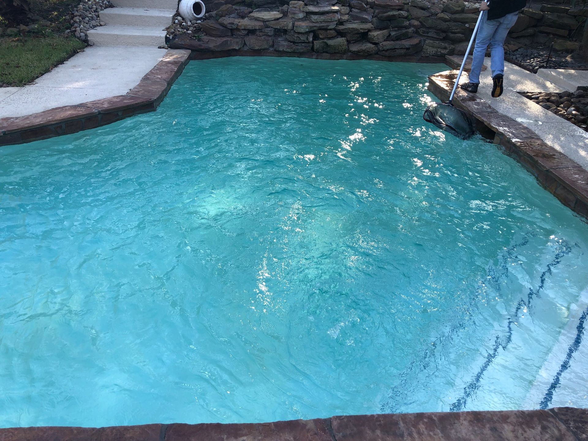 Blue backyard swimming pool with stone edges and a person standing at the right side
