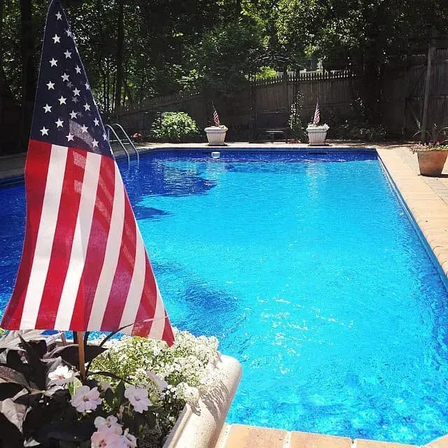 American flag by a bright blue backyard pool with white flowers and trees in the background