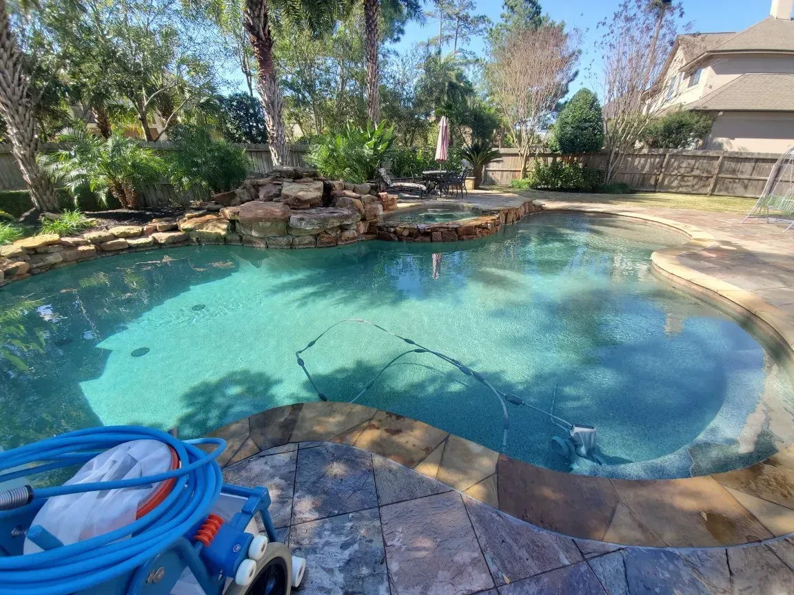 Backyard swimming pool with turquoise water, stone edging, trees, and a blue pool cleaner in the foreground