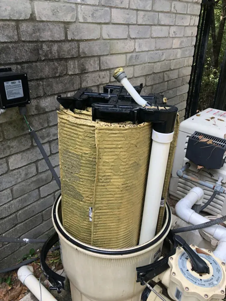 Damaged yellow filter canister with torn top, beside white pipes and equipment outdoors by a brick wall