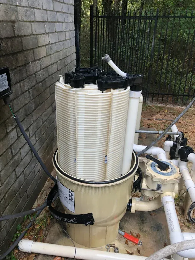 Large beige cylindrical water filter tank with white cartridge filters and pipes outdoors beside a brick wall and fence