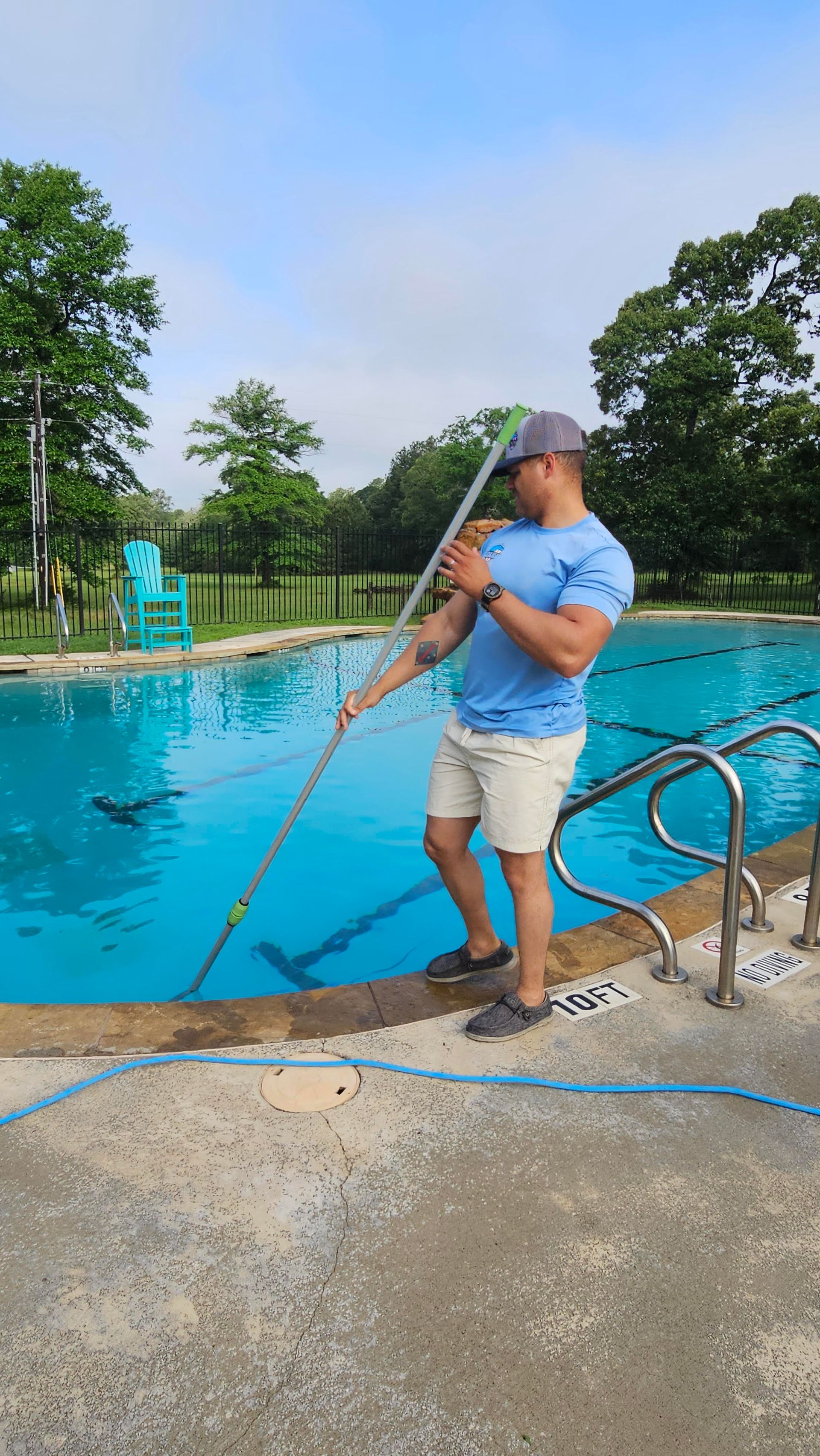 White dog standing in a shallow pool under a tan umbrella, outdoors by a wooden fence and trees