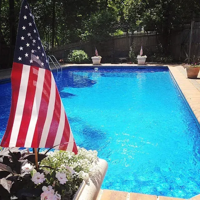 American flag beside a bright blue backyard pool with potted plants and flowers