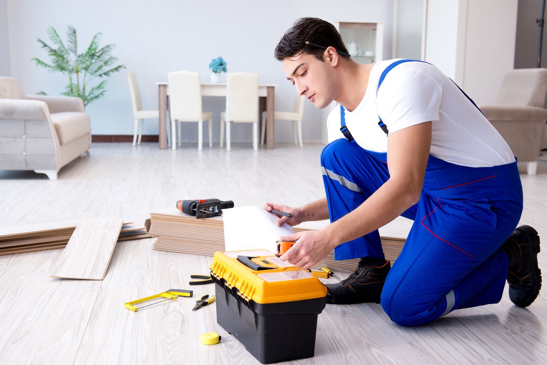 A person kneels, looking at a toolbox, wood planks nearby. They wear blue overalls in a bright living room.