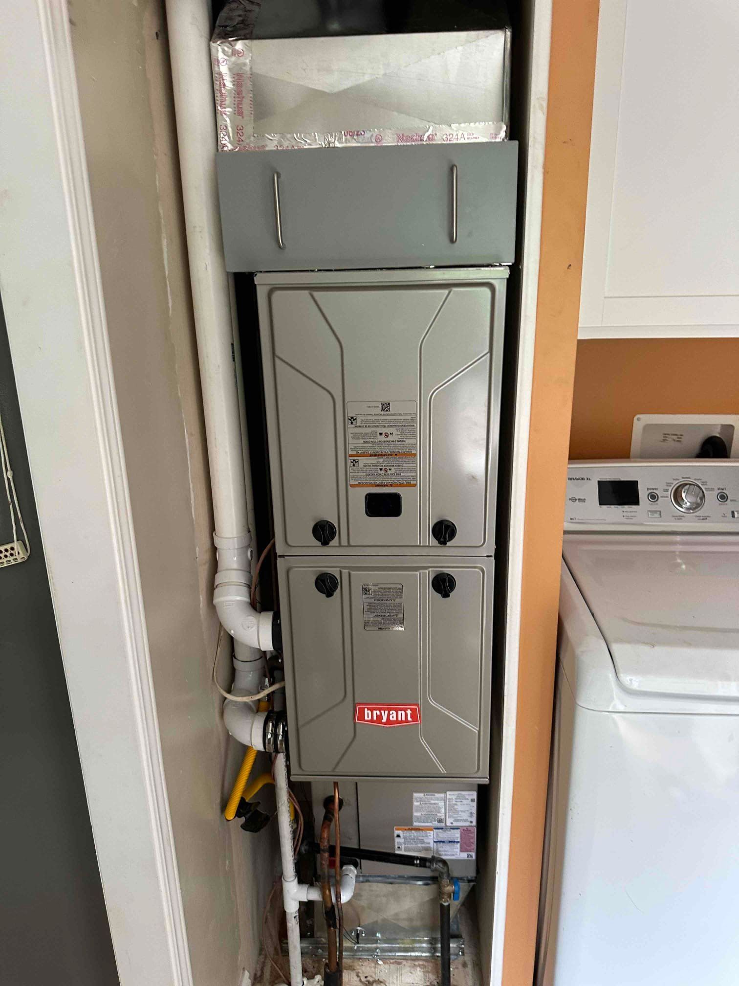 Home HVAC furnace installed in a utility closet beside a washer, with exposed ducts and pipes.