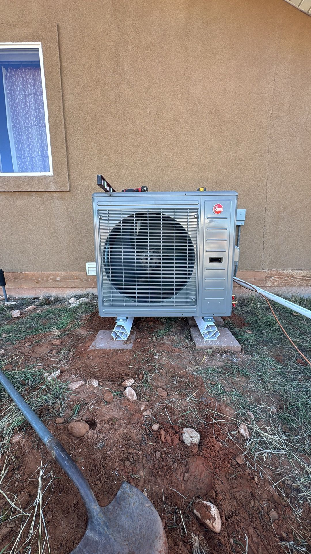 Outdoor air conditioning unit beside a tan stucco wall and window, on dirt ground with hoses nearby