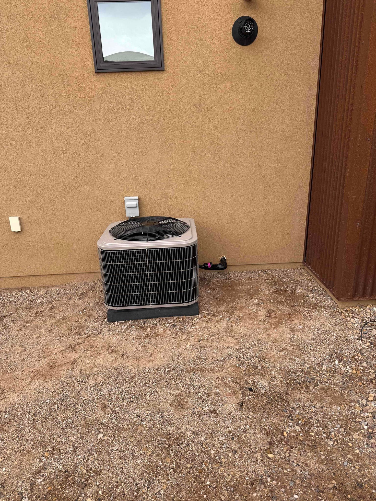 Outdoor air conditioner unit against a tan stucco wall and gravel ground, with a window above.