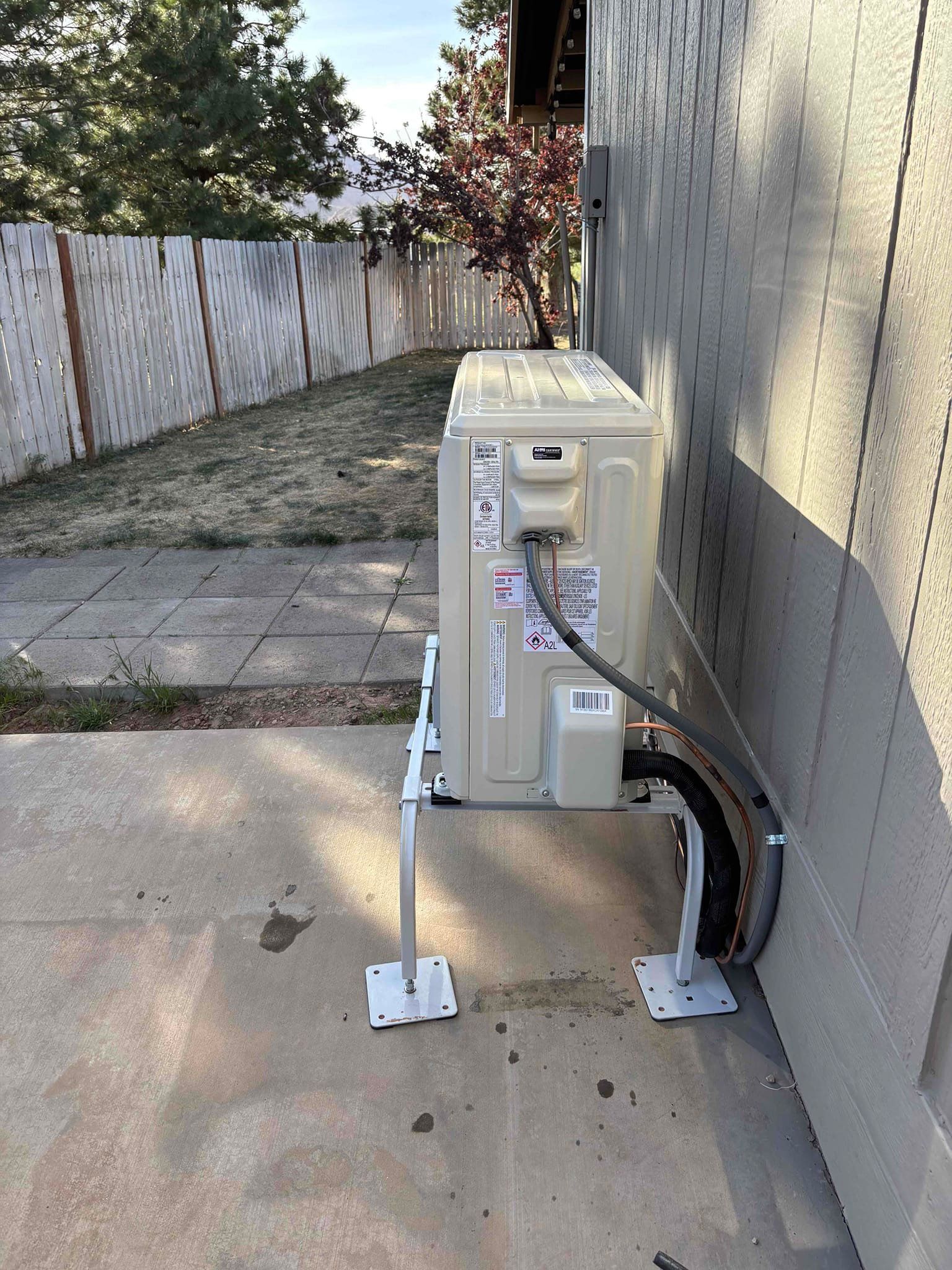 Outdoor HVAC unit beside a house, with a concrete pad and fenced yard in the background