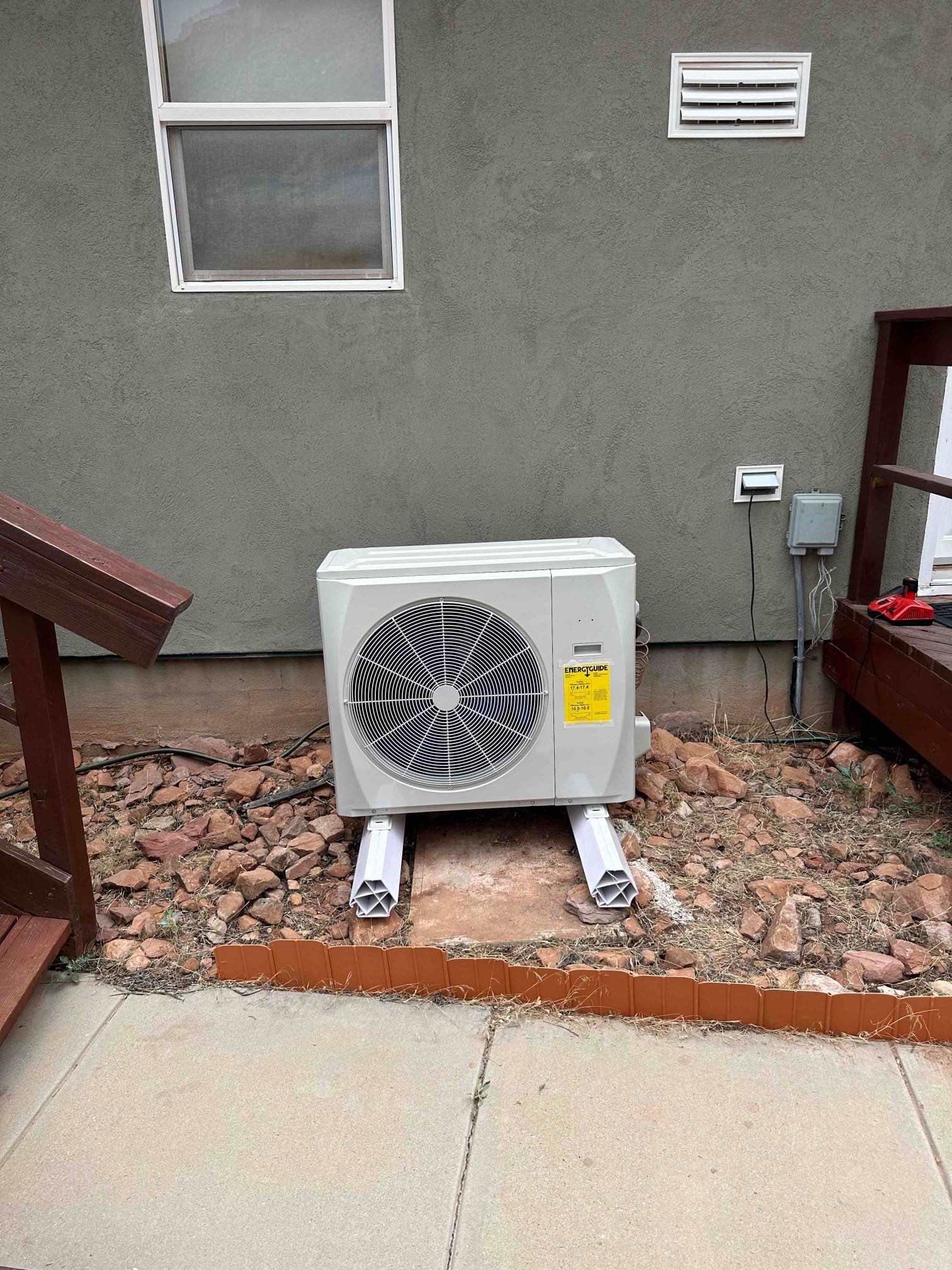 Outdoor AC condenser unit on gravel beside a house wall, with a window and vent above it.