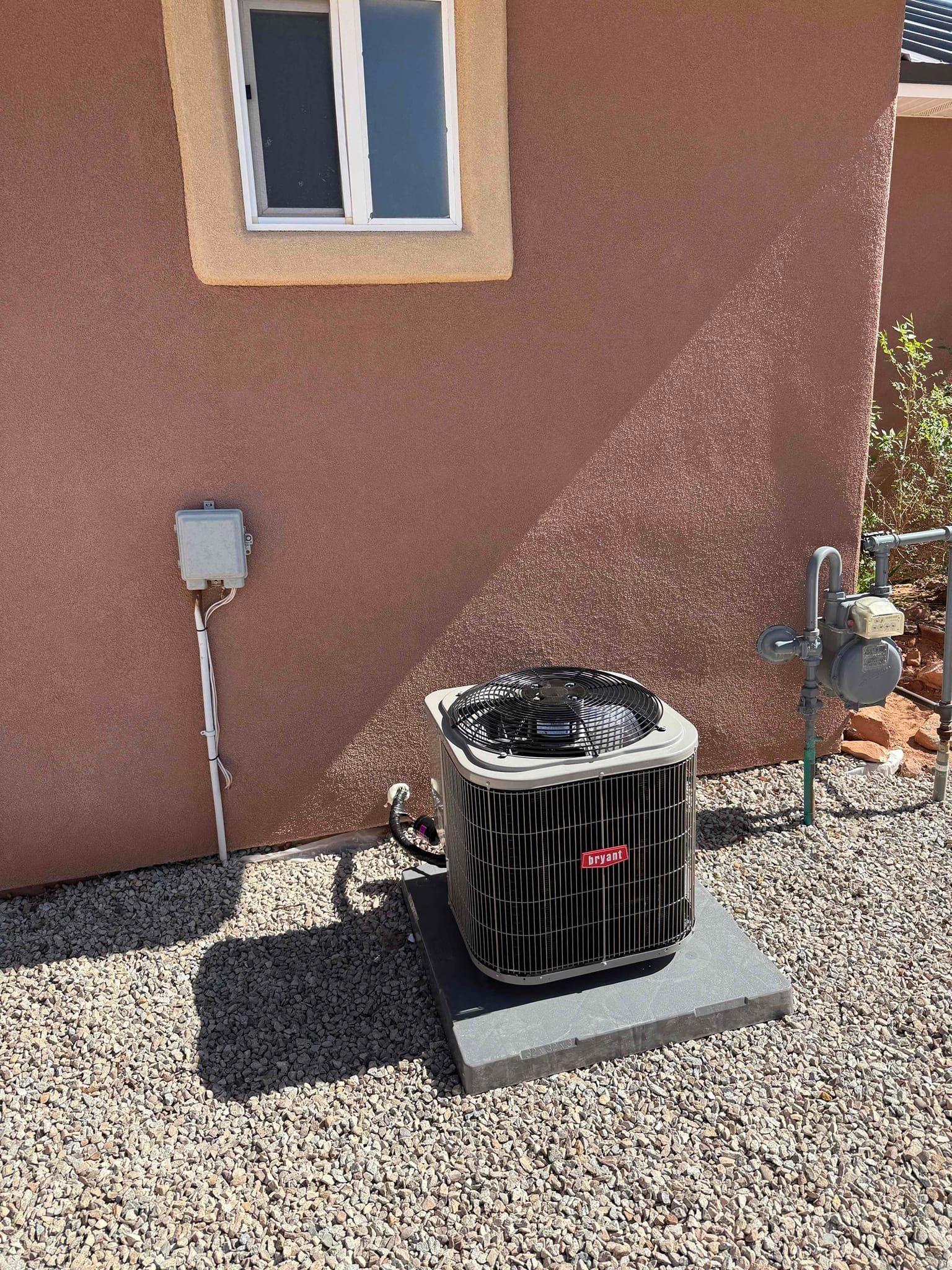 Outdoor HVAC unit beside a stucco wall under a window, on a gravel yard.
