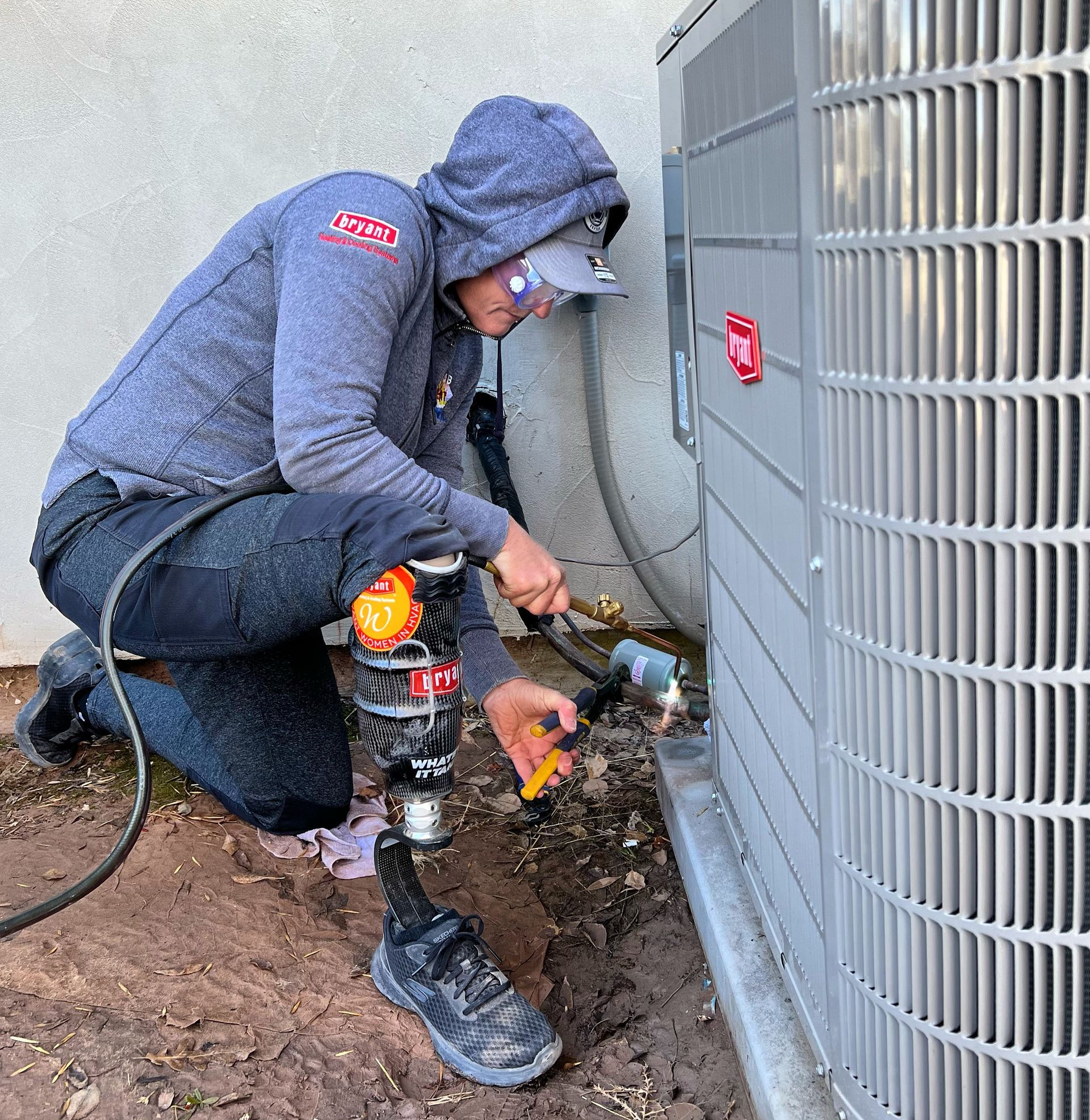 Worker crouches beside an outdoor HVAC unit, using tools on piping near the ground.