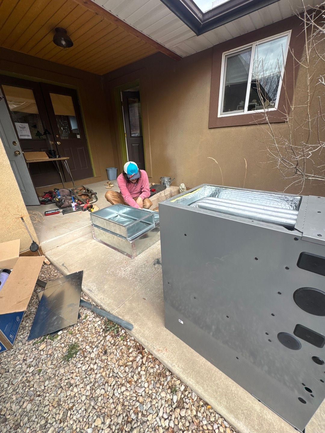 Person working beside HVAC units on a covered patio, with tools and boxes nearby.